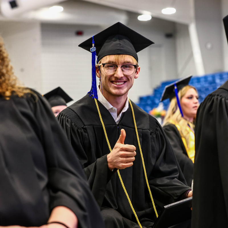 Student in cap and gown smiling at the camera holding up a thumbs up